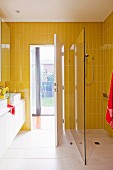 Gelbe Fliesen in Badezimmer mit Waschtisch und bodengleicher Dusche mit Glasabtrennung; Blick durch geöffnete Tür in den Garten