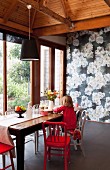Red and white chairs at rustic wooden table, little girl kneeling on chair, floor-to-ceiling terrace doors and floral wallpaper on back wall
