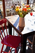 Poppies of various colours in china jug in sunshine on vintage table with red-painted kitchen chair to one side