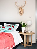 Round oak side table next to upholstered bed with floral scatter cushions below stylised antlers on wall