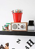 Coloured pencils in red pot in top of radio flanked by miniature chair and toys on black, wall-mounted shelf