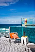 Holiday atmosphere - cane chairs and white stools on wooden deck, pool with transparent walls and view of Pacific Ocean in background