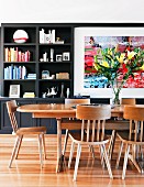 Oak dining table and bouquet of lilies in front of dark living room shelving with integrated painting