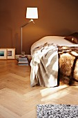 Modern, designer standard lamp and stack of books next to bed with fur blanket against brown-painted wall