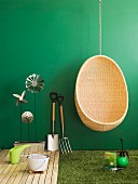 Wicker hanging chair next to gardening utensils and garden ornaments arranged on wooden decking and artificial lawn against green wall