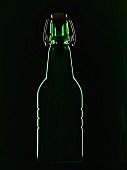 A green beer bottle with a flip-top lid backlit against a black background