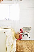 Vintage rattan chair next to bed with floral blanket against white, wooden wall and window with airy curtain