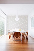 White dining table, classic wooden chairs, patterned wallpaper on end wall and glass wall to one side in minimalist room
