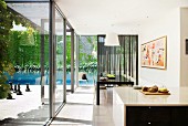 Open-plan kitchen and dining area next to continuous glass wall; view of sunny terrace and pool with climber-covered back wall