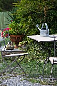 Garden table with watering can and garden chair in front of bush and planted urn
