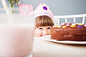 A little girl wearing a crown hiding behind a decorated chocolate cake