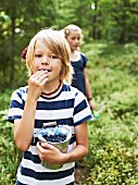 A little boy in a forest eating freshly picked blueberries with a little girl in the background