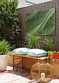 Wicker lantern on terracotta floor in front of bamboo bench with cushions and photo print on screen fence in courtyard