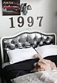 Double bed with curved headboard upholstered in silver; ornamental numbers and retro lamp on wall