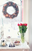 Wreath with fairy lights hung in window, gnome figurine and vase of red tulips on white vintage tray