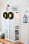 Christmas wreaths of fir branches and ribbons hanging on cupboard door in rustic interior with white wood cladding