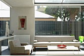 Sitzbereich in einem Glasvorbau mit hellen Designer Polstermöbeln und farbigen Glasgefässen auf Beistelltischen; Sammlerstücke in Wandnische und seitlicher Konsole