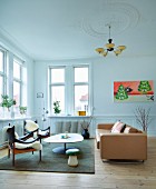 Wood-framed armchairs with white fur blankets, pale brown leather sofa and coffee table with amorphous top in living room with Art Deco pendant lamp and stucco ceiling