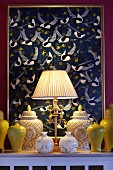 Table lamp with pale, pleated lampshade, collection of yellow ceramic vases and lidded jars in yellow and white in front of artwork with pattern of birds