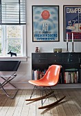 Charles Eames rocking chair with orange shell seat in front of floating sideboard below framed posters