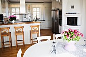 Bouquet of tulips on white dining table, counter and bar stools in front of white, open-plan country-house kitchen