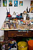 Paints and paintbrushes on vintage work table below postcards of artworks pinned to wall of painter's studio