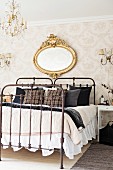 Vintage, twin beds with wrought iron frames below antique, oval, gilt-framed mirror on wallpapered wall