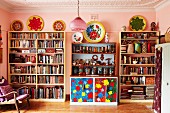 Library area with pink-painted walls and colourful floral dresser between full bookcases in collector's apartment