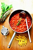 Bolognese sauce in a pan on a wooden board with penne pasta, herbs and olive oil