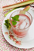 A glass of rhubarb spritzer with ice cubes and pieces of rhubarb on an old, rose patterned plate