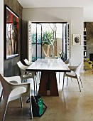 Massivholztisch und weiße Designer Stühle auf hellem Estrichboden, im Hintergrund offene Terrassen Flügeltür und Blick auf Palme im Patio
