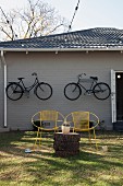 Yellow, vintage wire chairs and tree stump table in front of grey wooden house with bicycles hung on facade