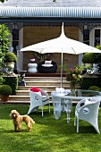 White wicker chairs and glass table below parasol in front of veranda steps with dog on lawn in foreground