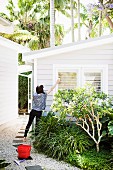 Frau beim Fensterputzen - Weisses Holzhäuschen mit kleinem Vorgarten in tropischer Umgebung