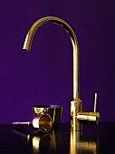Brass, designer tap fitting and brass-coloured utensils (bell and bowl) against violet background