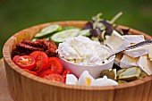 A wooden bowl filled with various salad ingredients (tomatoes, cucumber, goat's cheese and Brie)