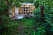 Overgrown path made from sleepers in garden outside house with lattice window and weathered facade