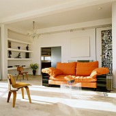 Sofa with orange upholstery and set of plexiglass coffee tables in living room; wooden designer chair in foreground