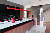 Long kitchen counter with wooden base cabinets below pendant lamp with red-painted narrow housing in modern kitchen with staircase in background