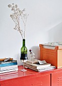 Vase aus einem Weinglas und einer abgeschnittenen Flasche, zusammen mit Bücherstapel und Akten auf rotem Metallspind