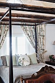 Scatter cushions arranged on dark, wooden four-poster bed below window in bedroom with wood-beamed ceiling