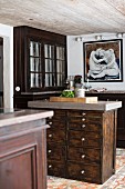 Free-standing chest of drawers made from dark wood with pale worksurface in front of old glass-fronted cabinet in rustic kitchen
