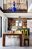 Solid wooden table and matching bench on concrete floor in front of kitchen area with yellow bar stools below mezzanine in open-plan interior