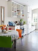 Helle lichtdurchflutete Küche mit pflegeleichtem Zementboden, offenem Küchenregal, rundem Esstisch mit farbigen Stühlen und Sitzbank am Fenster mit Stauraumschubladen
