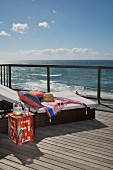 Comfortable sun lounger with cushions and painted side table on wooden deck; view of ocean over balustrade