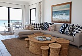 Rattan coffee table with matching stools of various sizes and modern corner sofa in open-plan interior; dining area in front of panoramic window in background