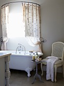 White chair and side table next to vintage bathtub below shower curtain on semi-circular curtain rail