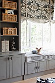 Custom, fitted, grey-painted cabinets, shelving and window seat with small dog sitting on cushion below Roman blind with ornamental pattern