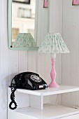 Black, vintage telephone and table lamp with pink base on small, white cabinet below wall-mounted mirror