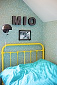 Yellow, vintage metal bed frame, pale blue and white polka-dot bed linen, charcoal wall lamp and decorative letters on patterned wallpaper in corner of teenager's bedroom